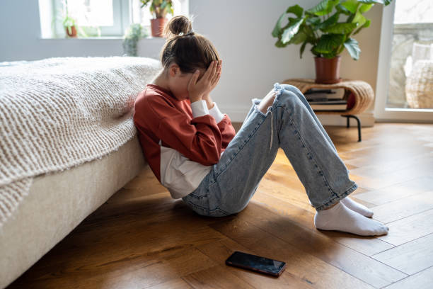adolescente malheureuse se couvrant le visage avec des mains pleurant alors qu’elle est assise sur le sol avec un téléphone portable à proximité - crying girl photos et images de collection