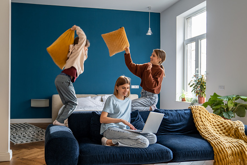 Naughty brother and sister freaking out playing pillow fight near busy smiling mom with laptop sitting on home sofa. Laughing kid boy and older sister indulge together distract mother resting on couch