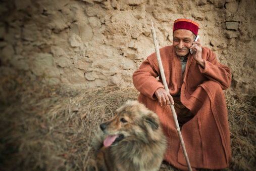 Real people from rural India: Senior Man using mobile phone, he is sitting against adobe wall holding a stick with his happy pet dog, he is in traditional dress of Himachal Pradesh.