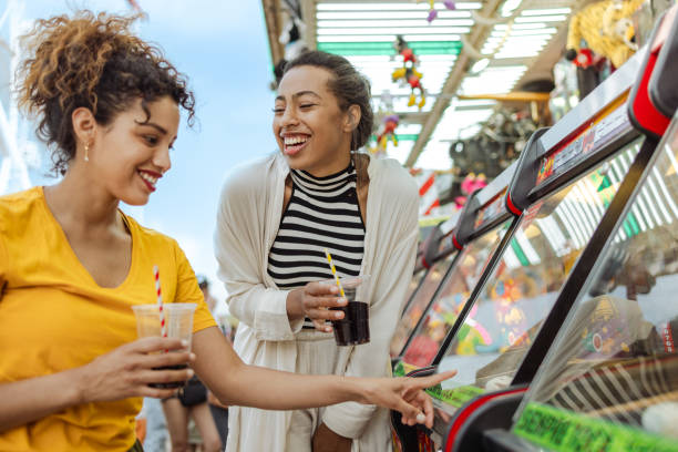 Playful female friends spending the day at the fun fair together. They are standing by the game machine and drinking.
