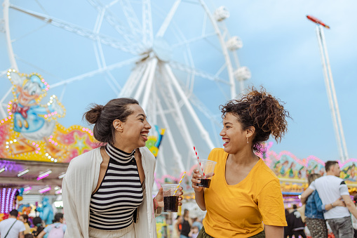 Portrait of happy friends having fun in the amusement park. They are talking, laughing and drinking. Behind them is a Ferries Wheel.