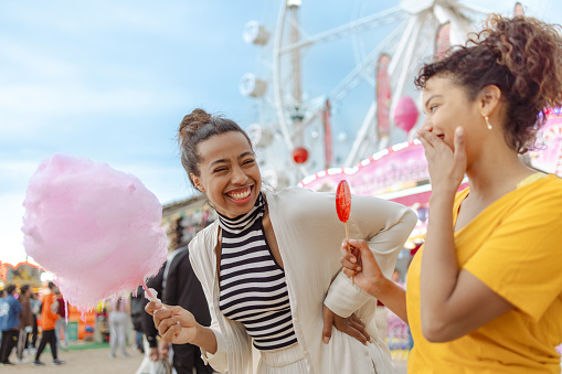 Portrait of cheerful female tourists in casual clothes relaxing and enjoying a Carnival. They are laughing and eating candies. Happiness is cheap.