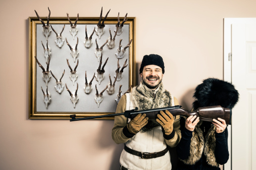 Funny portrait of hunter and his wife. They are standing near hunting trophies - antlers - and holding gun. Studio shot, home interior.