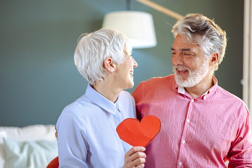 A senior couple is at home, they are embracing while holding a heart-shaped symbol