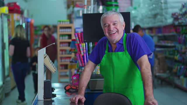 Happy senior staff of supermarket standing at cashier checkout wearing green apron and smiling at camera