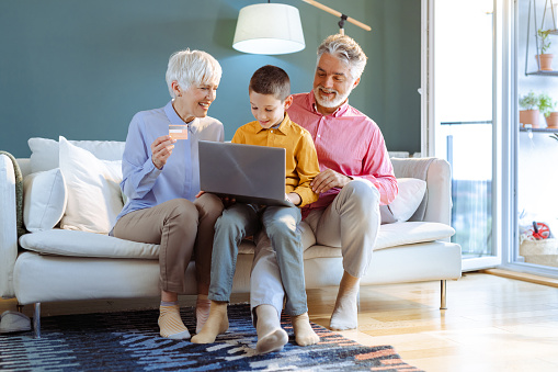 Shot of grandparents sitting on the sofa in the living room with their grandchild. They are smiling and using online paying services