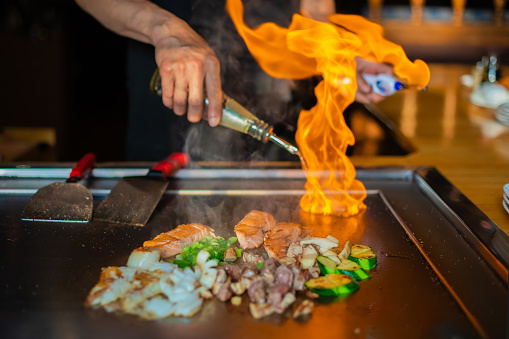 chef's hands with spatula over teppanyaki. cooking vegetables meat and seafood on hot hibachi grill table. Traditional Japanese Cuisine. Teppan show with flambe