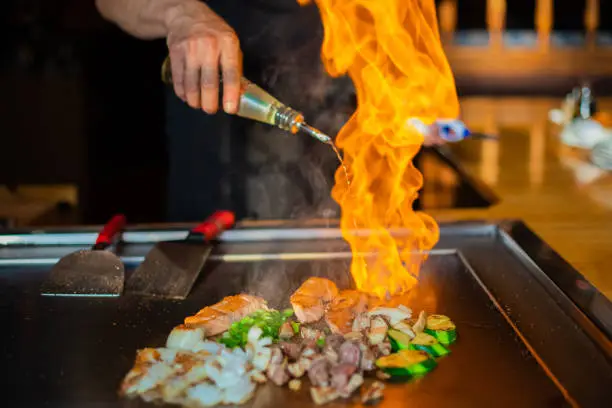 chef's hands with spatula over teppanyaki. cooking vegetables meat and seafood on hot hibachi grill table. Traditional Japanese Cuisine. Teppan show with flambe