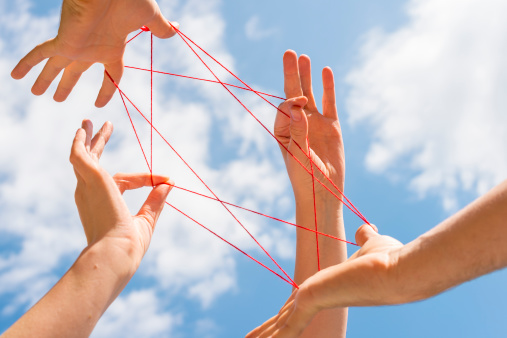 Four hands (female and male) against blue sky playing cats cradle with red wool.