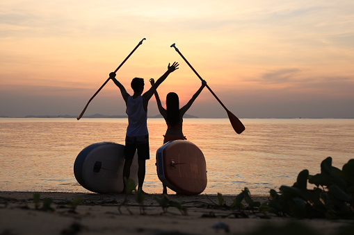 Silhouette of man and woman paddleboarding at sea, recreation sport paddling ocean beach surf. Young fit couple support each other, raising hands with paddles