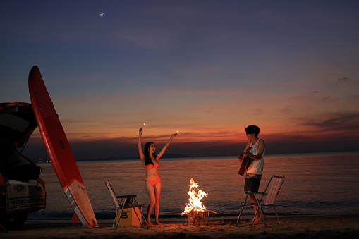 A couple Enjoying A Campfire by a young man playing guitar and a young girl holding a sparkler firework and Dancing On The Beach at Sunset.