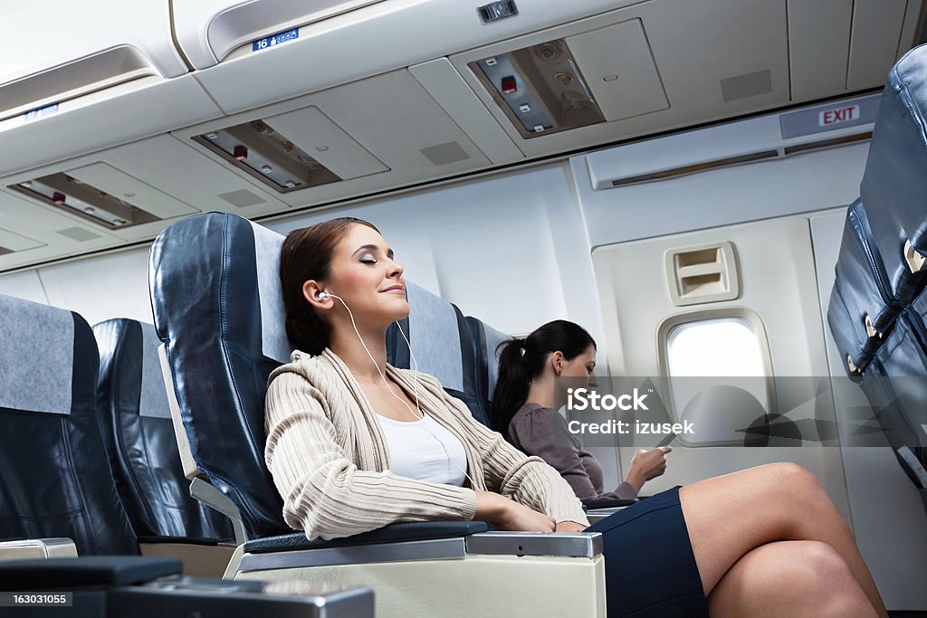 During the flight Young woman sitting on the airplane with earphone and listening to the music, while her neighbor reading a book. Airplane Stock Photo