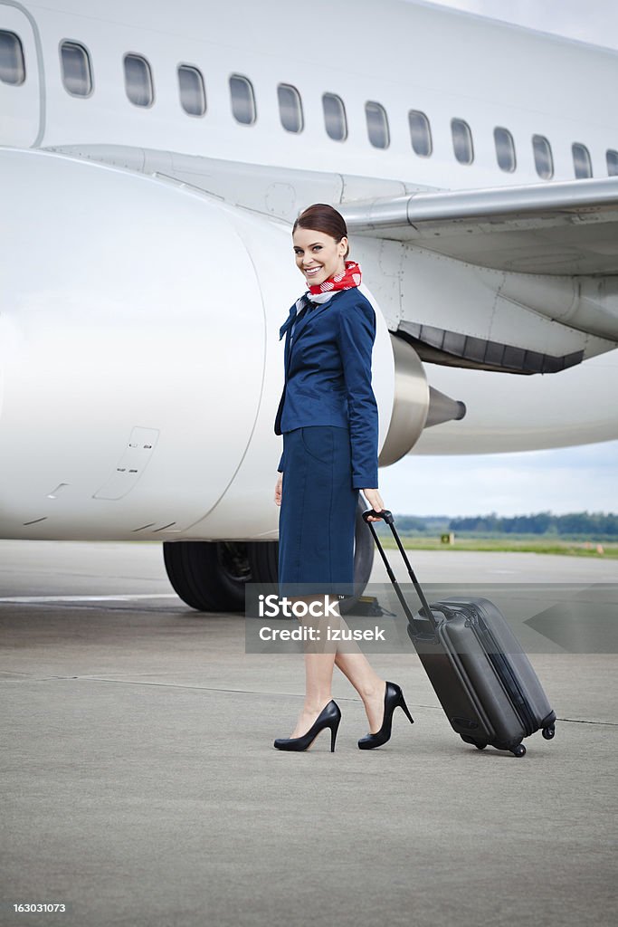 Air stewardess Outdoor portrait of a beautiful flight attendant walking with suitcase with an airplane inthe background, smiling at the camera. Cabin Crew Stock Photo