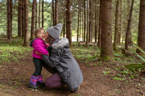 Eurasian pregnant mother of Asian and Hawaiian descent kneels next to her two year old daughter and gives the child a kissing on the cheek. The family is hiking through the forest while on a camping trip in the Pacific Northwest region of the United States.