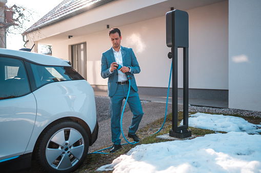 Full length shot of a mature asian man standing next to his electric car. He is holding the charging cable from its charging station.