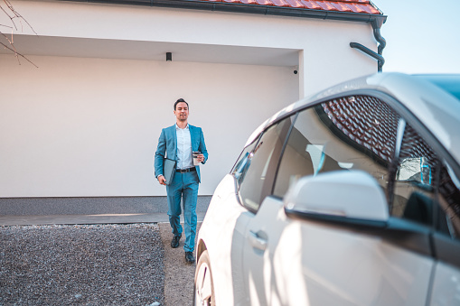 Morning outside of a modern home. Japanese mature man is walking to his electric car to get to work on time.