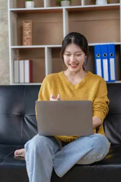 Photo of Beautiful young Asian woman in casual clothes Use a laptop and smile while sitting on the couch. work from home Work remotely, surf the net, New Normal concept