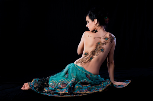 Low key profile portrait of beautiful, brunette woman sitting on the ground with her back to the camera. She has a peacock feather henna design on her back. Shot in the studio on a black background.