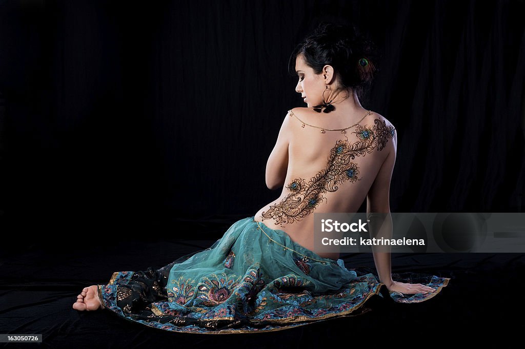 Peacock Feather Henna Design on a Woman’s Back Low key profile portrait of beautiful, brunette woman sitting on the ground with her back to the camera. She has a peacock feather henna design on her back. Shot in the studio on a black background. Back Stock Photo
