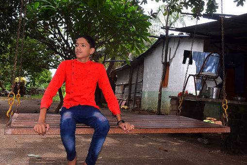 Indian boy in red shirt and blue jeans is sitting in an iron swing. garden background