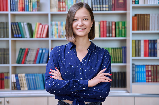 Portrait of confident female college student with crossed arms looking at camera, inside library in educational building. Knowledge, education, youth, college university concept