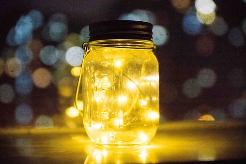 A glass jar containing yellow lights atop a table illuminated against a bokeh background