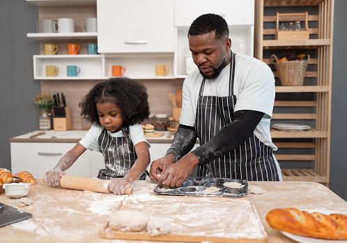 Happy African American kid girl and father cooking break or bakery with rolling bread in kitchen at home