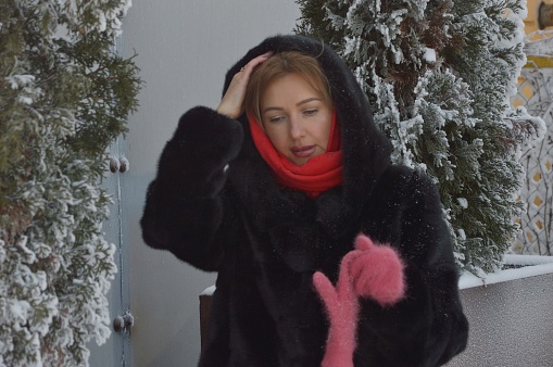 Young woman in a cozy fur coat with a hood and pink mittens on the street in winter. Portrait of a girl dressed in winter clothes.