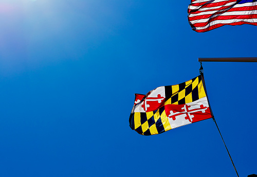 The Maryland state flag and U.S. flag fly in a strong wind on a bright, sunny day against a clear blue background.