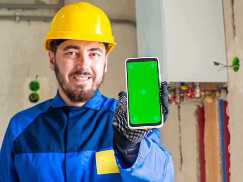 Portrait of worker or engineer in blue work clothes and yellow safety helmet with space for text. A man at a construction site holding a phone with chroma key screen in his hand and showing it.