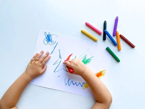 Photo of small child draws with pastel crayons on white table.