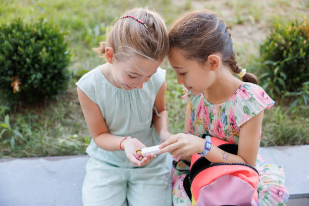 Two close friends elementary age girls sitting on surrounding wall on school grounds, sharing sweets on recess