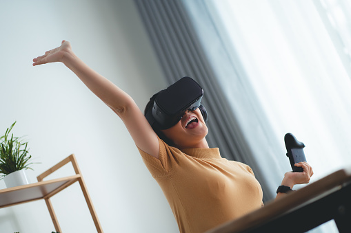 Asian woman in VR headset holding joystick happy and smiling while sitting at home office.
