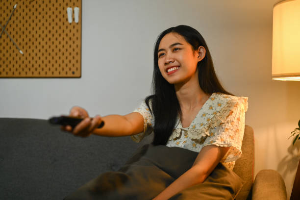 istock Happy asian woman watching television sitting on a comfortable sofa with the remote control in her hand 1631050848