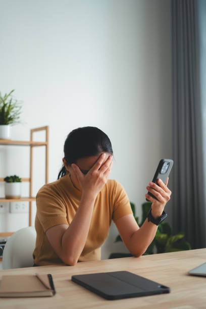 Asian woman sitting at home office looking at smartphone screen and having problem.