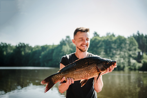Big carp. Happy fisherman hold big trophy fish near lake. Fresh fish trophy in hands. Young man returning with freshly caught fish. Article about fishing day. Fishing backgrounds. Success pike fishing