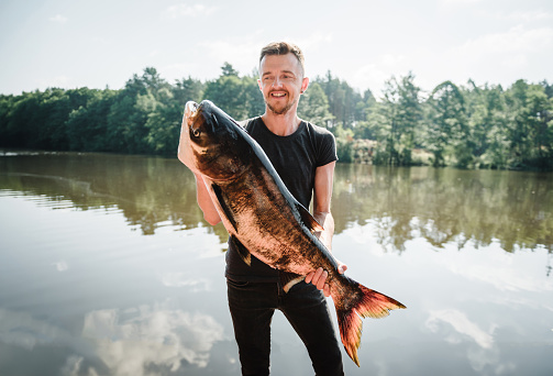 Happy fisherman hold big trophy fish near lake. Success pike fishing. Fresh fish trophy in hands. Young man returning with freshly caught fish. Article about fishing day. Closeup. Fishing backgrounds.