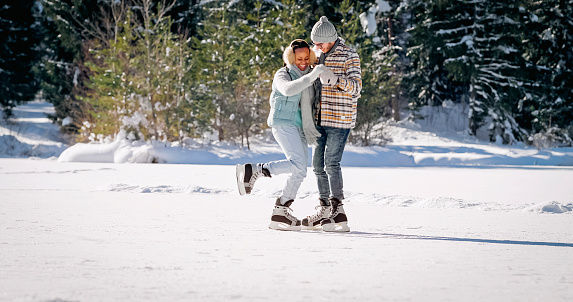 Smiling young couple holding hands while ice skating on frozen lake in winter.