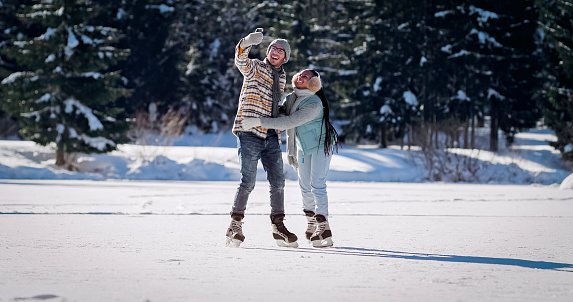 Smiling young couple taking selfie while ice skating on frozen lake in winter.