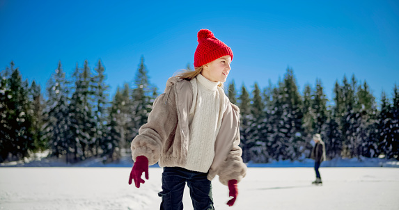 Girl looking away while ice skating against sky on frozen lake in winter.