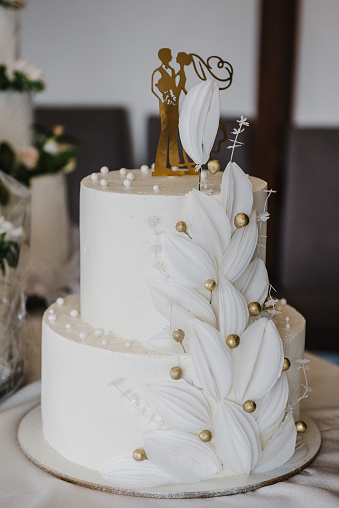 Wedding white cake decorated golden decor, figurines, topper of bride and groom suite and flowers for a banquet. Selective focus. Delicious reception for luxury wedding ceremony. Trendy cake. Closeup.