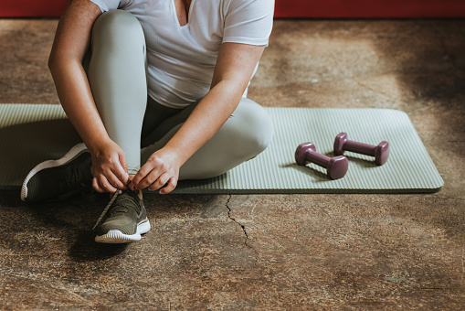 An anonymous plus size woman sitting on an exercise mat, tying her shoelace.