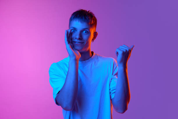 istock Excited smiling young man whispering secrets and pointing sideway against gradient pink purple background in neon light 1631159357