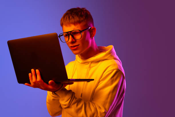istock Young man, student, freelancer in glasses attentively looking on laptop against gradient purple background in neon light 1631159919