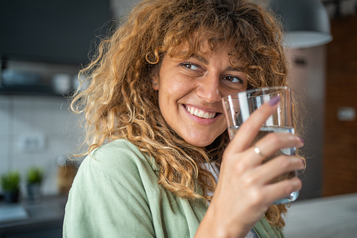 Portrait of a smiling young woman with curly hair holding a glass of fresh clean drinking water. Concept of healthy lifestyle and fluid intake