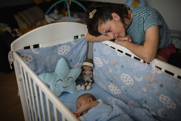 Cute silent little baby boy in white babysuite is in his crib, lying on his back, covered with blue blanket. He is sleeping while his mother taking care of him. Love emotion.