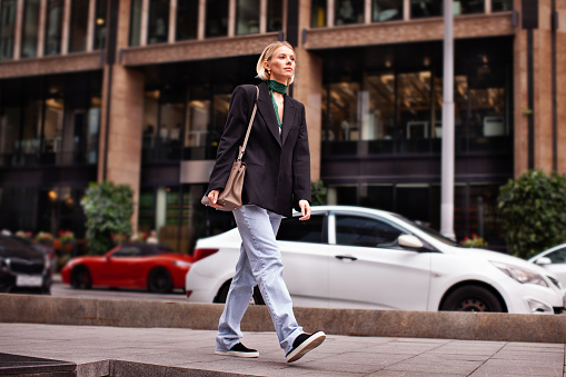 Beautiful young stylish woman walking down city street against backdrop of building, wearing fashionable clothes jeans, black blazer, handbag and sneakers. Fashion model serious and looks away.