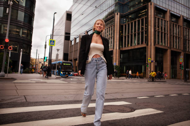 Beautiful and stylish young woman crossing road at pedestrian in city, smiling and dressed fashionably in jeans and blazer. Fashion street style on background of business center building stock photo