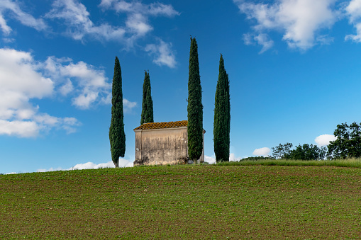 Ponsacco, PI, Italy-June 7, 2023; Low angle view of small church Chiesetta di San Pierino surrounded by Mediterranean Cypresses typical for Italian landscape against a white clouded blue sky