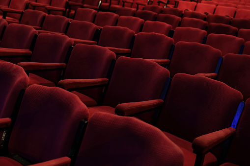 Empty rows of deep red fabric chairs or seats with arm rests in a theater or movie cinema hall.